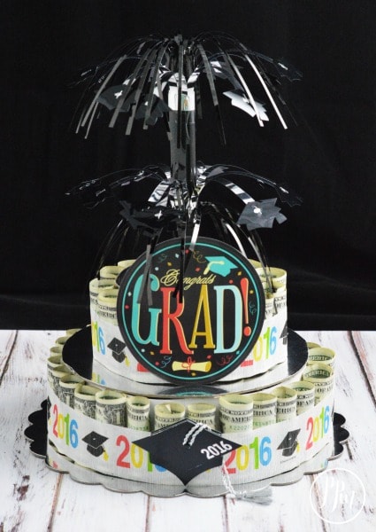 It looks like a two tiered cake. Each tier is made up of rolled up money and there is a round circle cut out that says "grad" and some black decoration protruding from the top that looks like fireworks