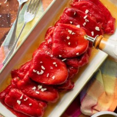 Roasted Red Peppers on cream colored platter with a drizzle of oil from dispenser and chopped garlic. Fork and spoon, multi colored napkin and dish of oil around.