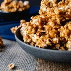 Homemade caramel corn in a grey bowl on blue cloth. smaller black bowl of caramel corn in background caramel corn and nuts on wooden table. close