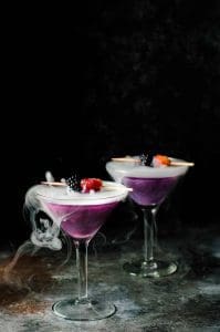 The Witches Heart cocktails in martini glasses on a dark counter. The liquid is purple