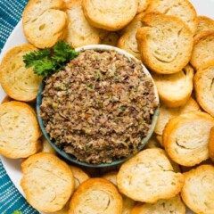 An overhead image of a bowl of Olive Tapenade on a large plate surrounded by toasted sliced baguette