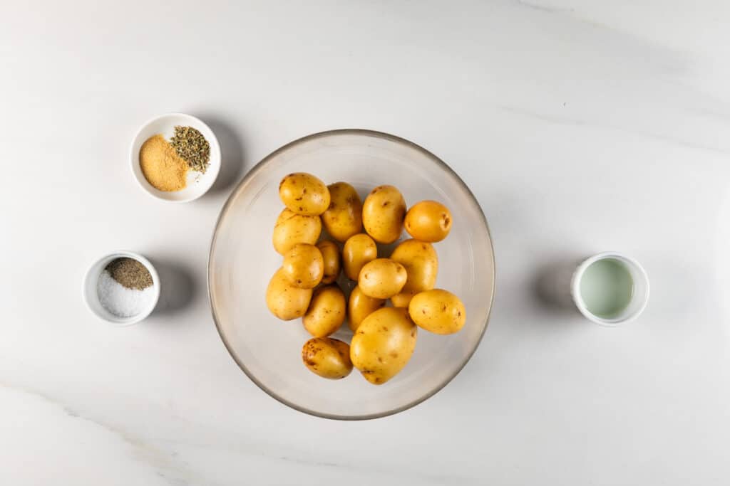 A bowl of small potatoes sits on a white surface, ready for the air fryer. Its surrounded by small bowls containing spices like garlic powder, herbs, salt, pepper, and a liquid, possibly oil. The ingredients are neatly arranged around the potatoes.