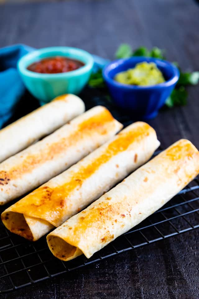 Close up shot of baked taquitos on cooling rack with dipping sauces in the background
