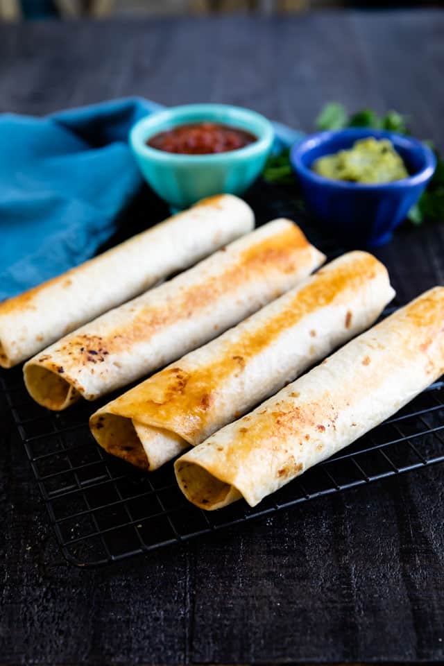 Four baked taquitos on a cooling rack with salsa and guacamole in background