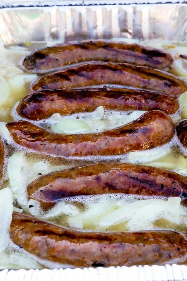 Close up shot of beer brats cooking in a deep metal pan with light beer and onions