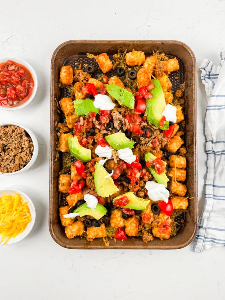 A tray of Tater Tot Nachos is piled high with black olives, avocado slices, diced tomatoes, sour cream, and ground meat. Small bowls on the side offer salsa, shredded cheese, and extra ground meat. A cloth napkin is placed nearby for a perfect party spread.