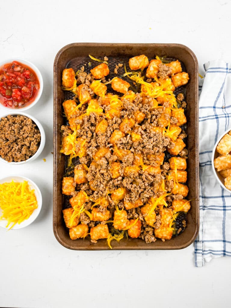 A baking tray showcases a tater tot casserole, reminiscent of delicious Tater Tot Nachos, topped with ground beef and melted cheese. Surrounding the tray are small bowls of salsa, shredded cheese, and extra ground beef. A blue and white checkered cloth lies to the side.