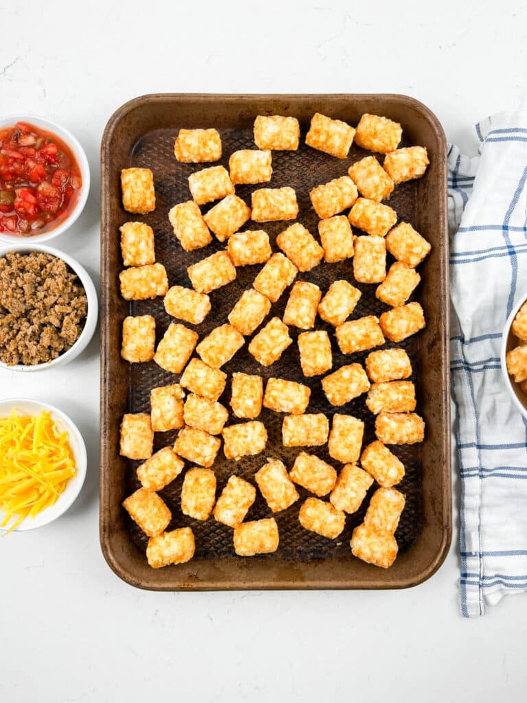 A baking tray loaded with golden tater tots rests on a white surface, ready for assembly into delicious Tater Tot Nachos. Surrounding the tray are small bowls of salsa, cooked ground meat, shredded cheese, and a dish towel nearby.