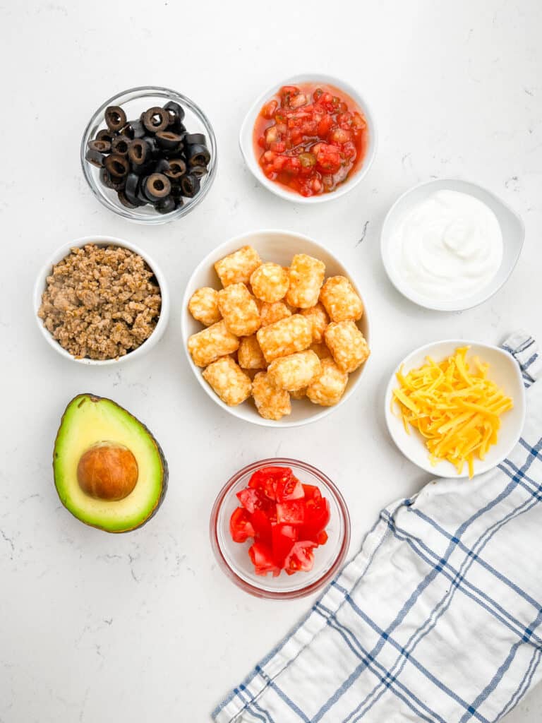 A flat lay of ingredients for Tater Tot Nachos on a white marble surface: tater tots, shredded cheddar, diced tomatoes, avocado, black olives, salsa, ground beef, and sour cream. A striped cloth napkin is placed at the bottom right.