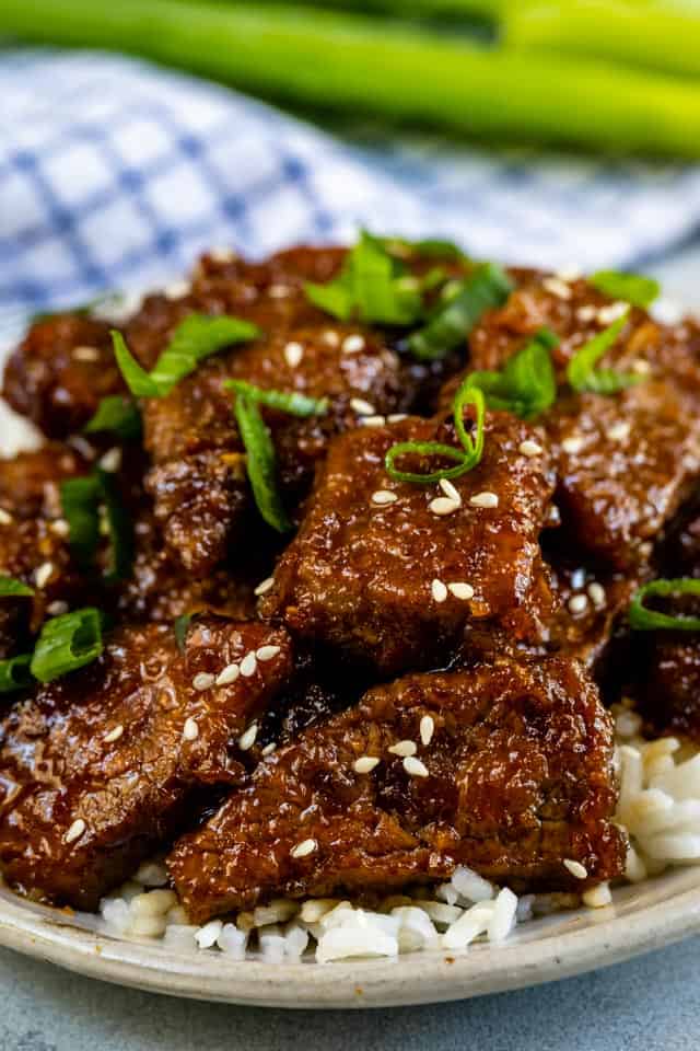 Mongolian beef with white rice on a grey plate with green onions and blue checkered napkin in background