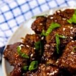 Overhead shot of mongolian beef on a grey plate with white rice and blue checkered napkin and green onions in background and recipe title on top of image