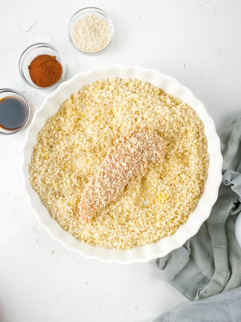 A raw chicken tender coated in breadcrumbs sits in a round white dish filled with more breadcrumbs—perfect for making crispy Air Fryer Chicken Tenders. Nearby are small bowls of spices, soy sauce, and extra breadcrumbs on a white countertop.