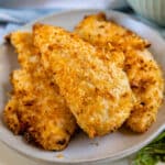 A plate of crispy Air Fryer Chicken Tenders coated in a golden breadcrumb crust is displayed, with a bowl of dipping sauce visible in the background.