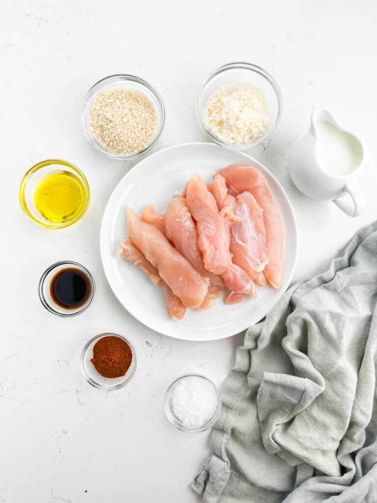 A white plate with raw Air Fryer Chicken Tenders is surrounded by small bowls of olive oil, panko breadcrumbs, grated cheese, soy sauce, paprika, salt, and a jug of milk on a white surface with a gray cloth.
