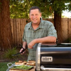 man in green shirt standing over bbq with chicken and asparagus on cutting board
