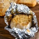 Roasted garlic wrapped in foil on a wood plate with garlic bread in background