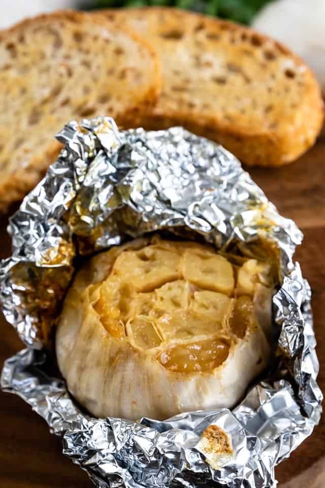 Close up of roasted garlic wrapped in foil on a wood plate with garlic bread in background