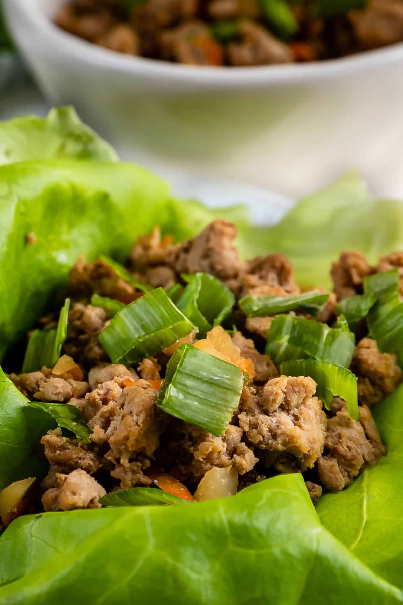Close-up of seasoned ground meat for Turkey Lettuce Wraps, topped with chopped green onions and served on fresh green lettuce leaves. A white bowl with more of the same filling is blurred in the background.