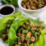 Two lettuce wraps filled with ground turkey on a plate with asian-style sauce and more ground turkey behind the plate