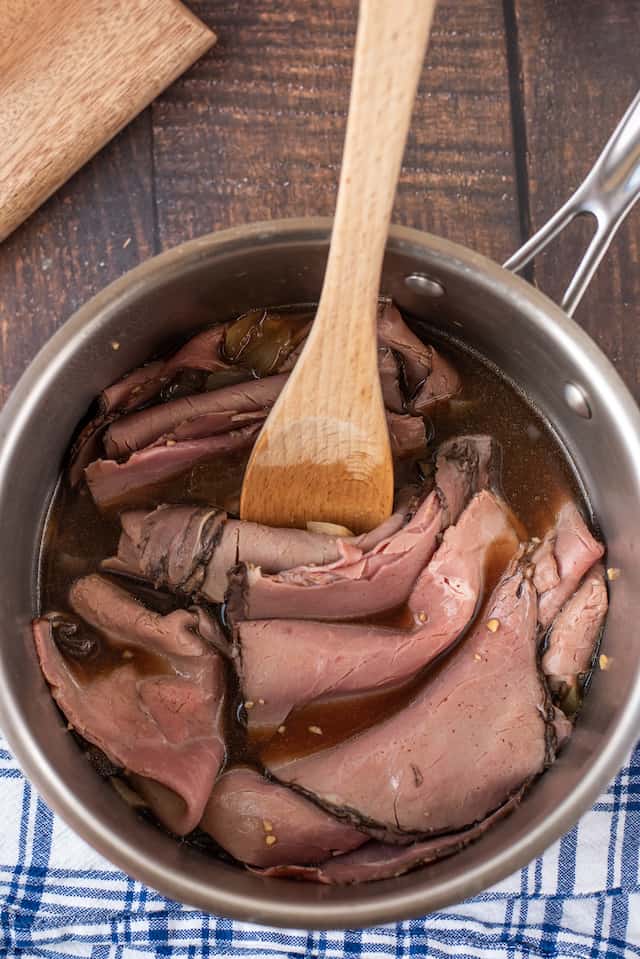 Overhead shot of deli meat and au jus in a saucepan with wooden spoon