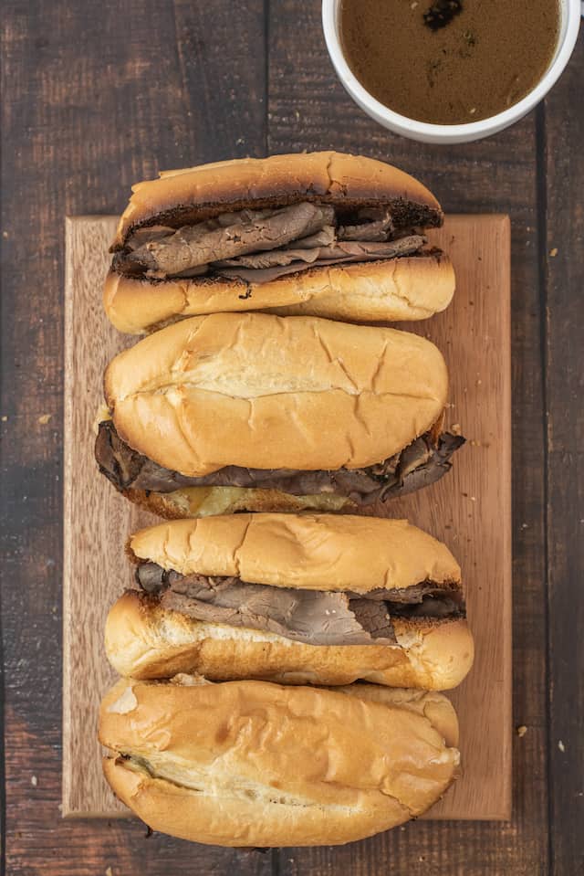Overhead shot of four french dip sandwiches on a wood cutting board with au jus on the side