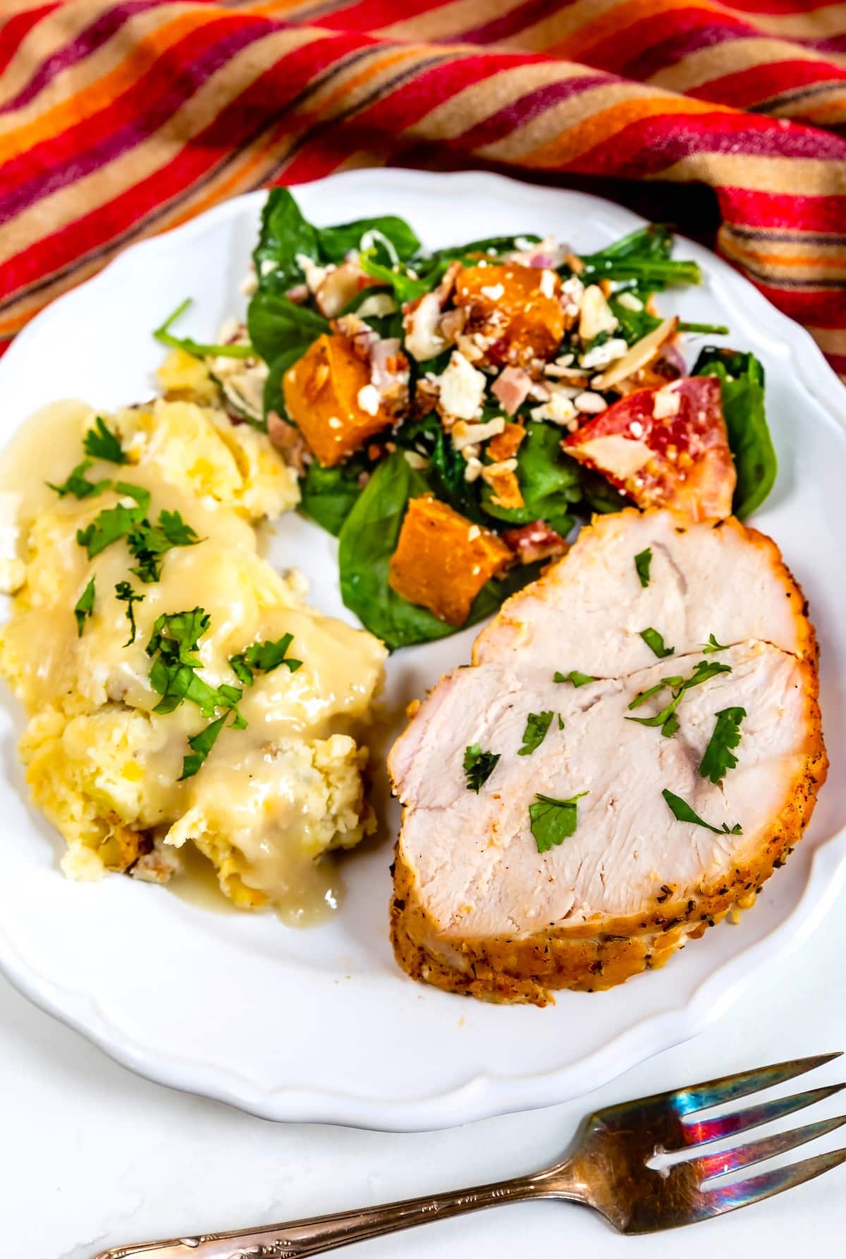 A white plate with sliced roasted turkey breast, mashed potatoes with gravy, and a spinach salad topped with sweet potatoes and feta cheese. A fork and a colorful striped napkin are nearby.