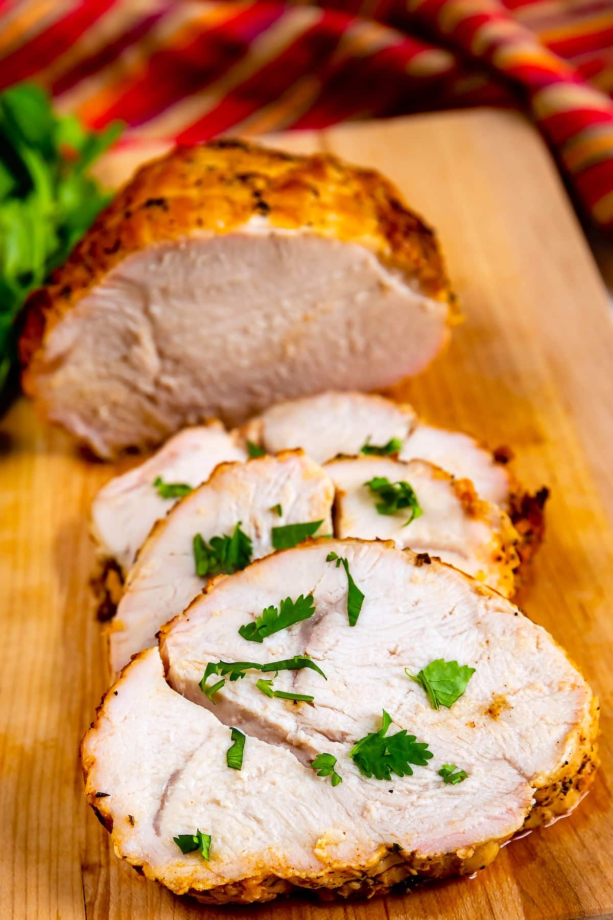 Sliced roasted turkey breast, featuring tender Roasted Turkey Breast, garnished with fresh parsley on a wooden cutting board, with a colorful striped cloth in the background.