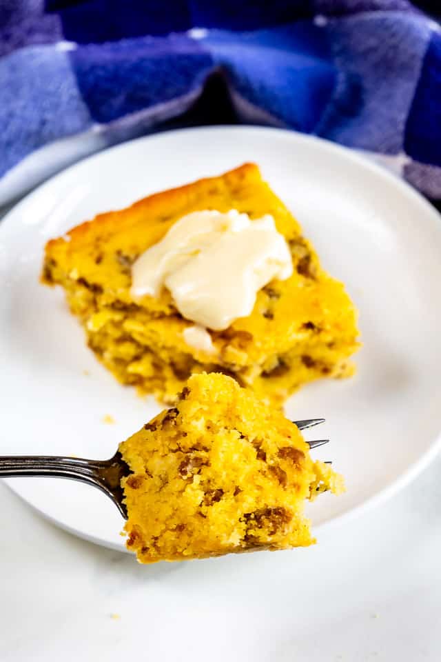 Slice of sausage cornbread with a pat of butter on top on white plate with one bite on fork