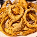 Close up shot of air fryer onion rings on a white plate with dipping sauce in background