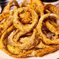 Close up shot of air fryer onion rings on a white plate with dipping sauce in background