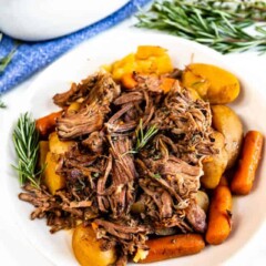 Crockpot pot roast on a white plate with gravy and herbs in background
