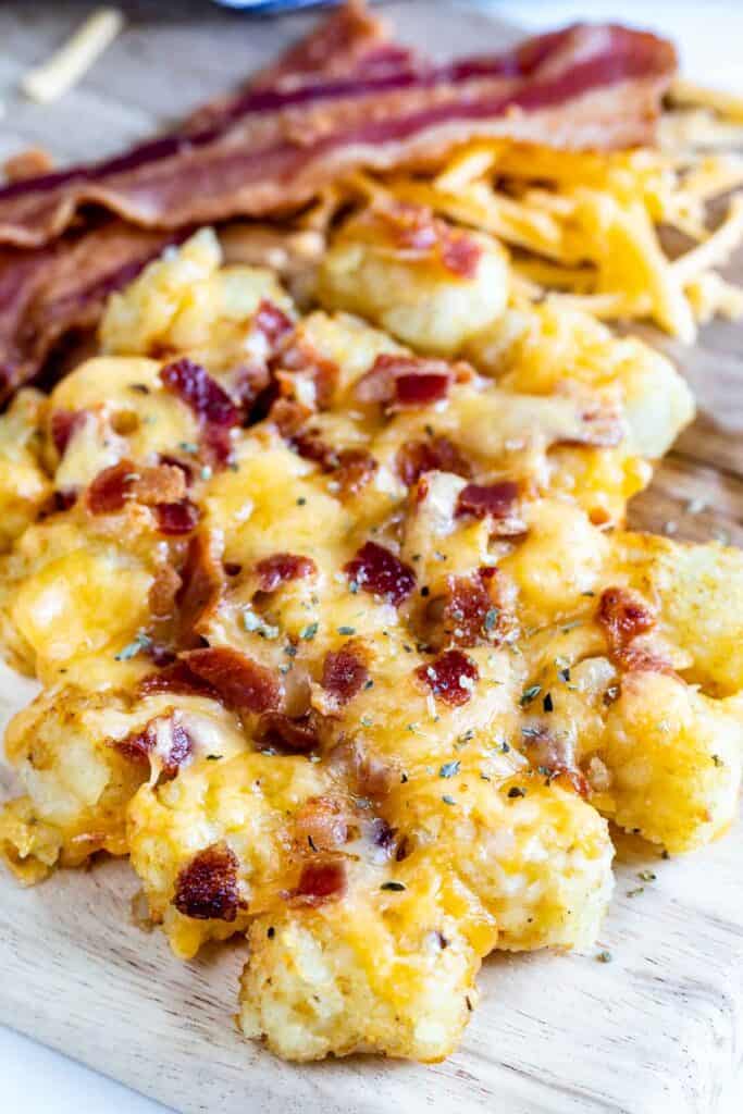 Loaded tater tots on cutting board with bacon strips behind them