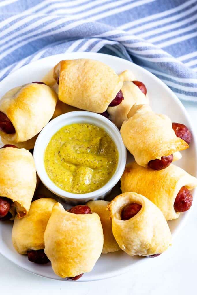 Overhead shot of pigs in a blanket in a circle on a plate with mustard dipping sauce in the middle