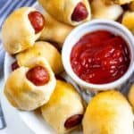 Overhead shot of pigs in a blanket in a circle on a plate with ketchup dipping sauce in the middle with recipe title on bottom left of photo