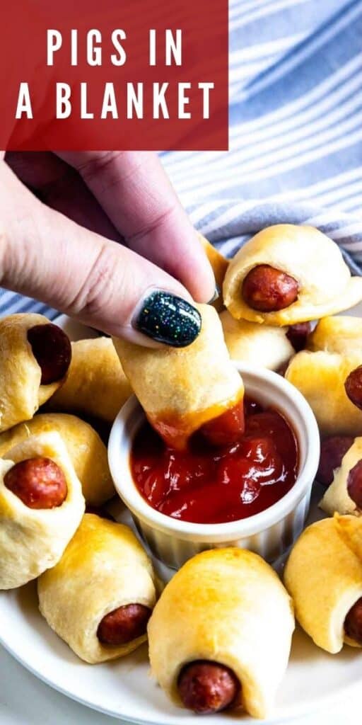 Hand dipping pig in a blanket into a dish of ketchup with recipe title on top right of photo