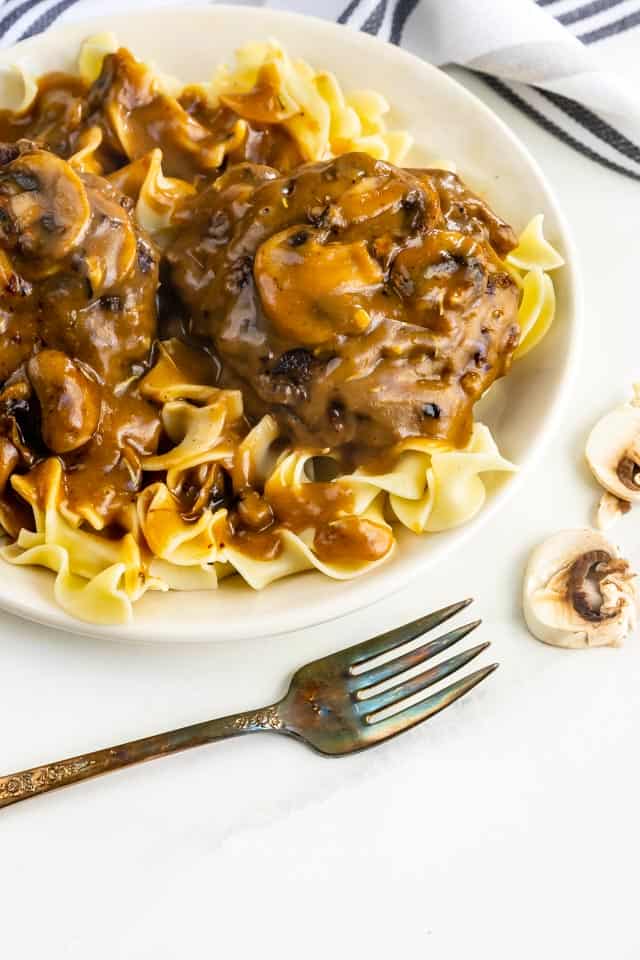 Overhead shot of salisbury steak over egg noodles on a white plate with fork and mushrooms next to plate