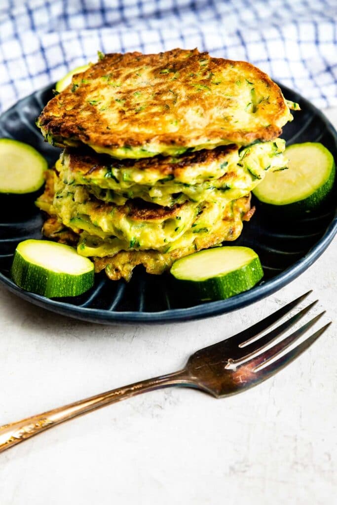 Stack of zucchini fritters on a plate surrounded by circled zucchinis