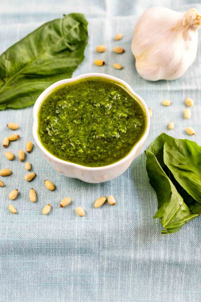 Basil pesto in a small white dish with garlic, toasted pine nuts and basil around it