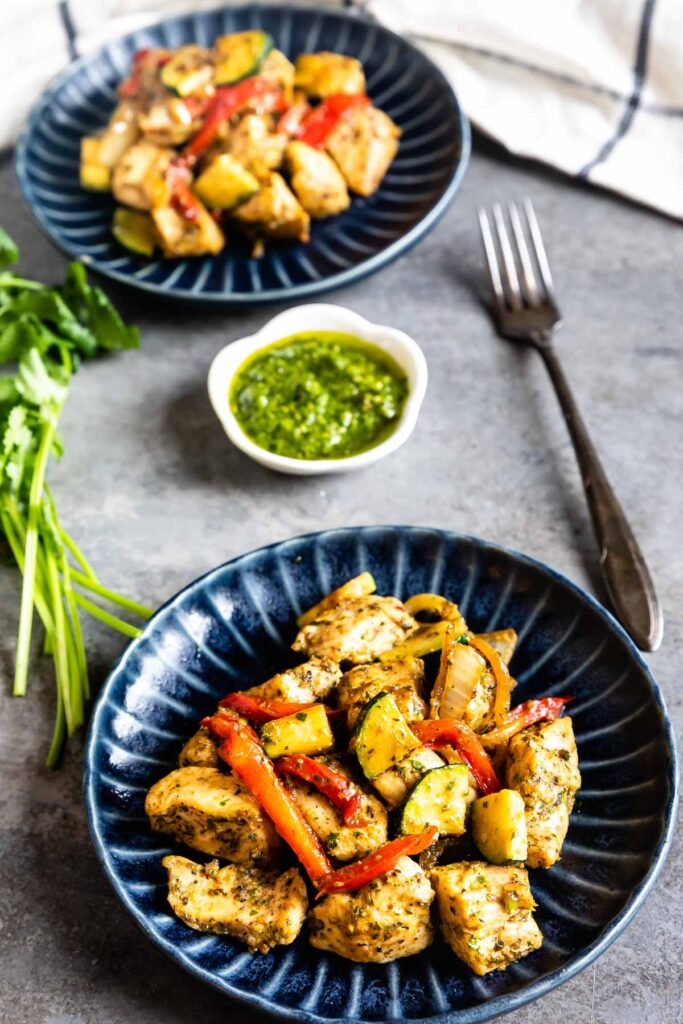 Two plates of pesto chicken and vegetables with a small dish of pesto in the middle