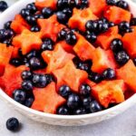 Big bowl of fruit salad with watermelon and dressing in background and recipe title on bottom of photo