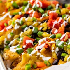Side shot of potato wedge nachos on sheet pan covered with toppings and guacamole in background