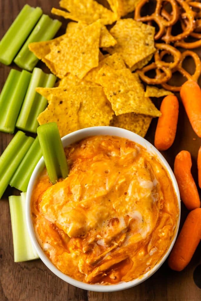 Overhead shot of buffalo chicken dip in a small bowl surrounded by chips, pretzels, carrots and celery