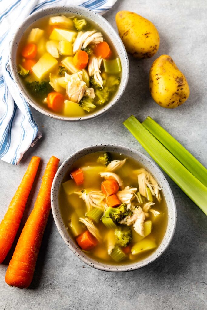 Overhead shot showing two bowls of chicken vegetable soup surrounded by carrots, celery and potatoes