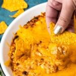Hand dipping a chip into the chili cheese dip with recipe title on top of image