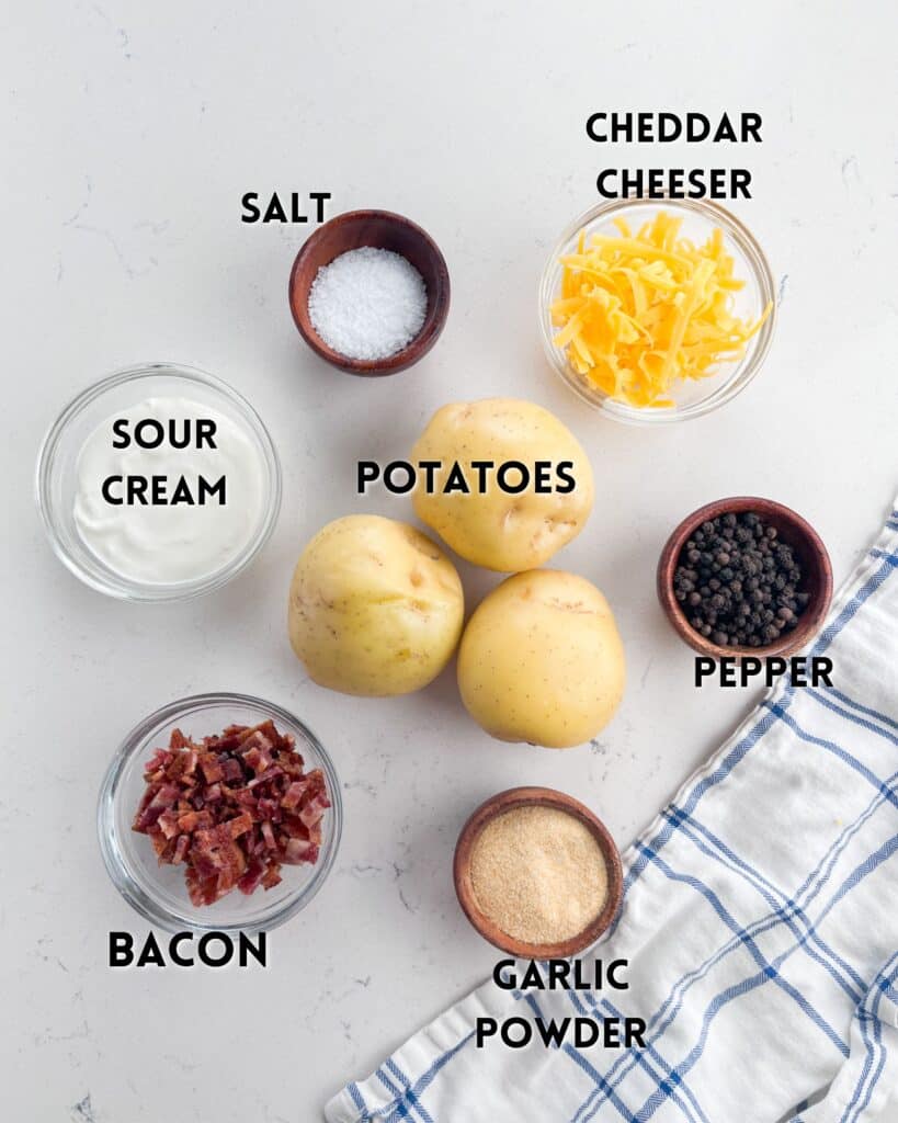 Overhead view of ingredients for Cheese and Bacon Smashed Potatoes, featuring three potatoes, sour cream, grated cheddar cheese, bacon bits, black pepper, garlic powder, and salt. A white cloth with blue stripes is placed nearby.