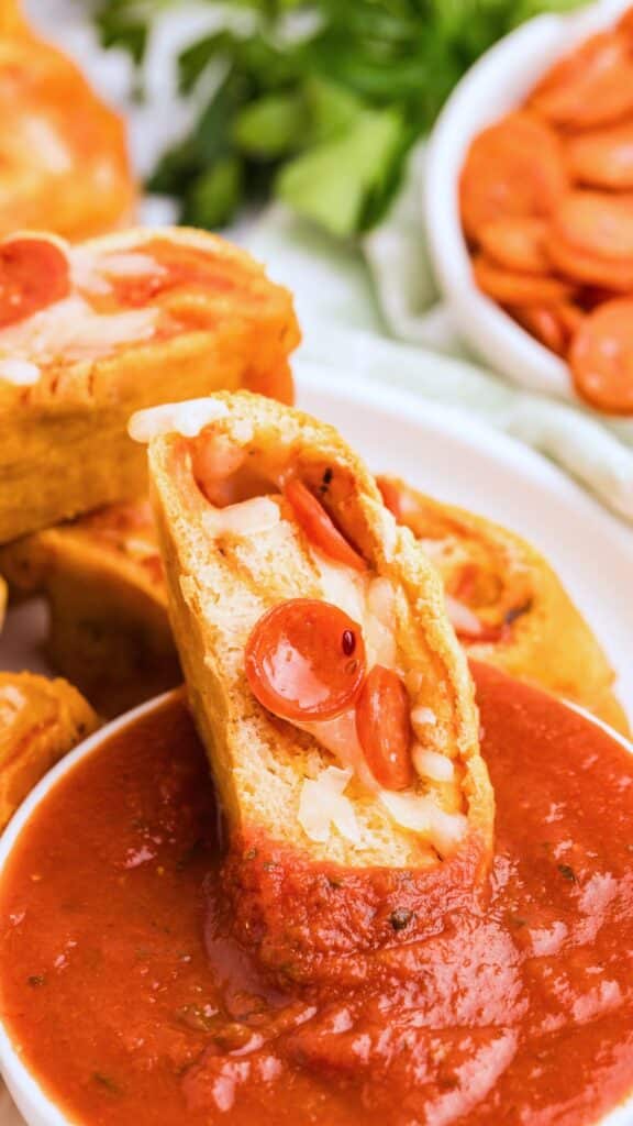 A close-up of a sliced bread roll akin to pizza rolls, stuffed with cheese and pepperoni, partially dipped in marinara sauce. A bowl of pepperoni slices and greenery are blurred in the background.
