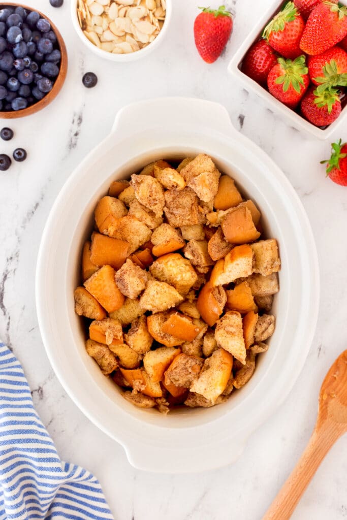 Overhead shot of french toast pieces in a white crockpot surrounded by optional toppings