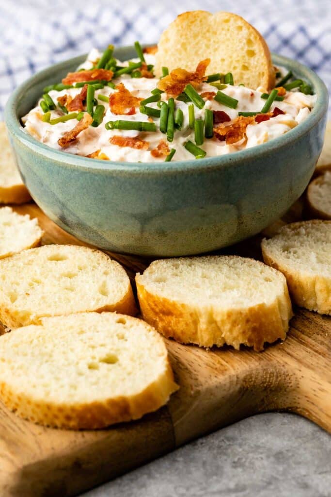 Picture of a bowl of baked potato dip with slices of baguette on a cutting board