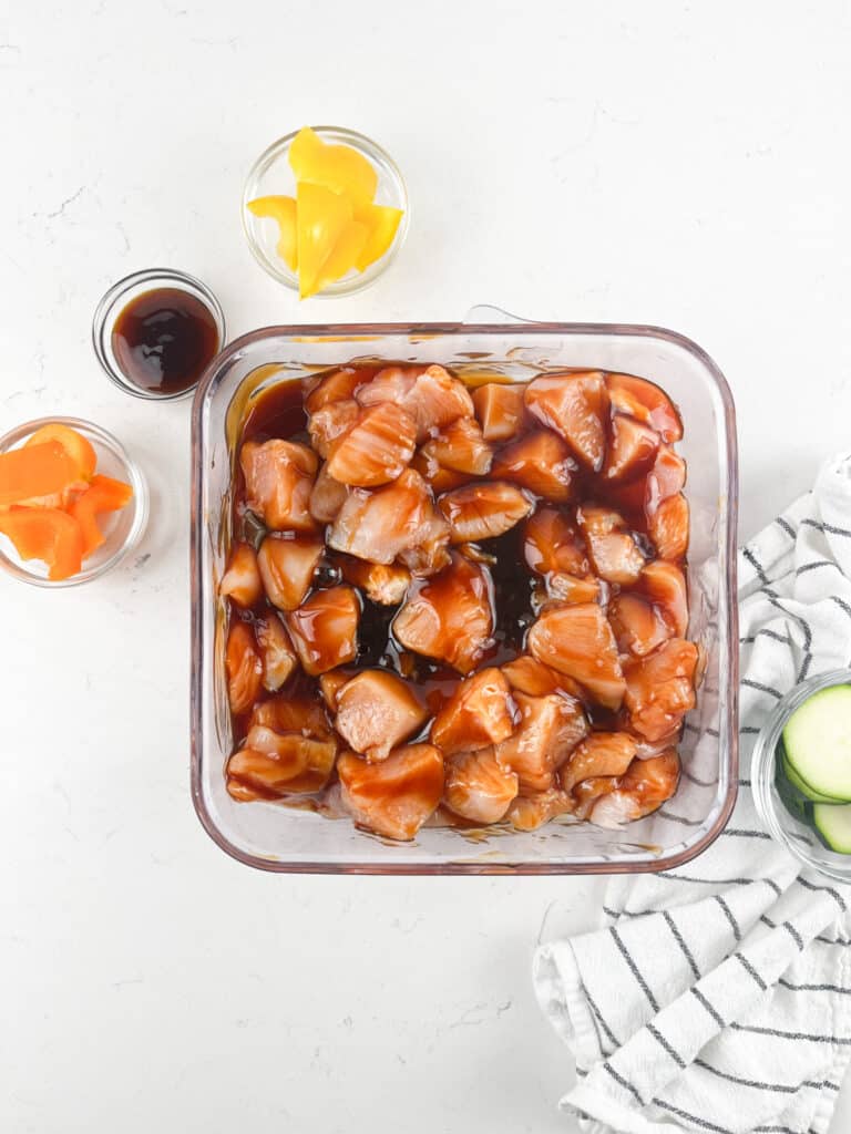 Overhead view of diced chicken marinating in a glass container with dark teriyaki sauce, perfect for chicken kabobs. Nearby, small bowls contain sliced yellow peppers, orange slices, and additional sauce. A striped cloth and cucumber slices are on the right. White background.