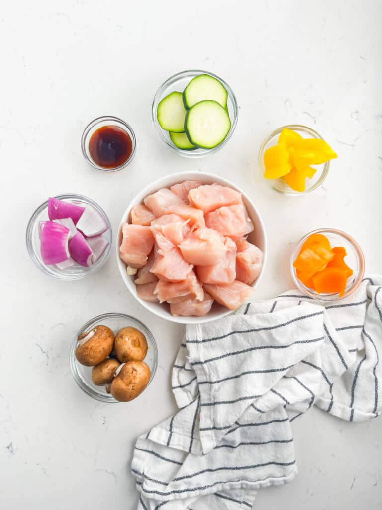 A variety of ingredients on a white surface: a bowl of raw chicken pieces in the center, destined for Teriyaki Chicken Kabobs, surrounded by small bowls of sliced zucchini, yellow and orange bell peppers, red onions, mushrooms, and a small bowl with sauce. A striped cloth is nearby.